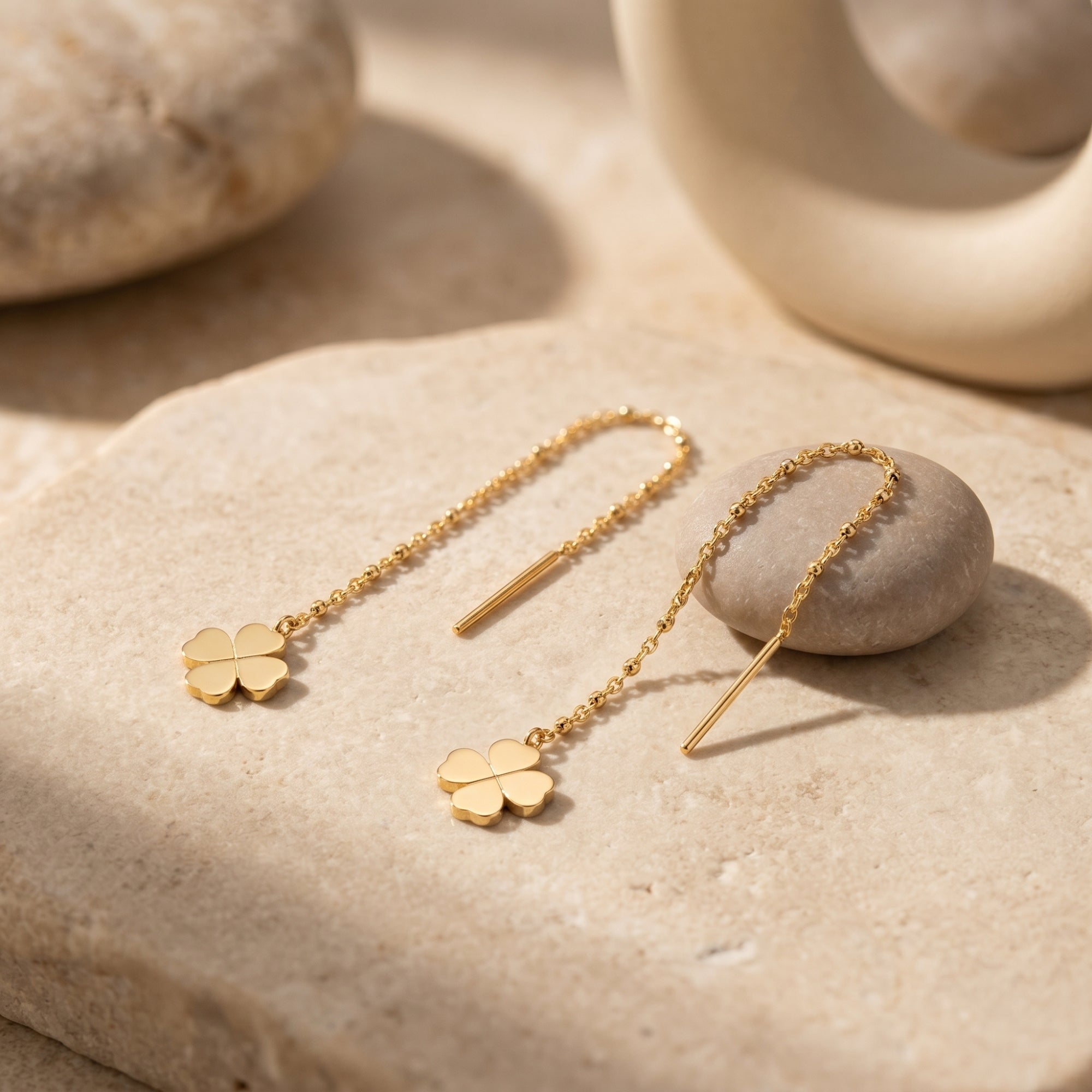 18K gold finish clover threader earrings displayed on a natural stone surface in warm sunlight.