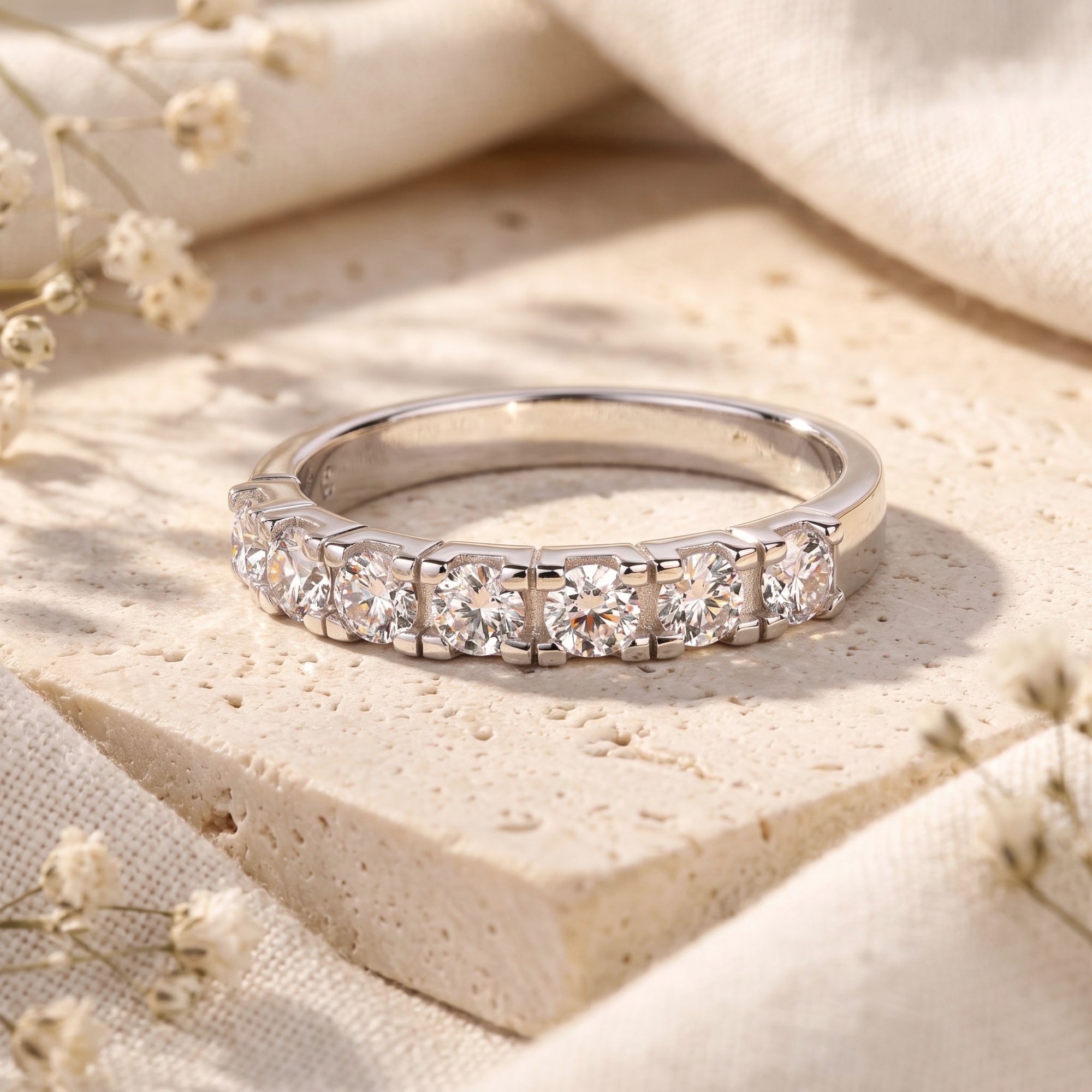 Silver half-eternity promise ring with round-cut simulated diamonds displayed on a stone surface with dried flowers.