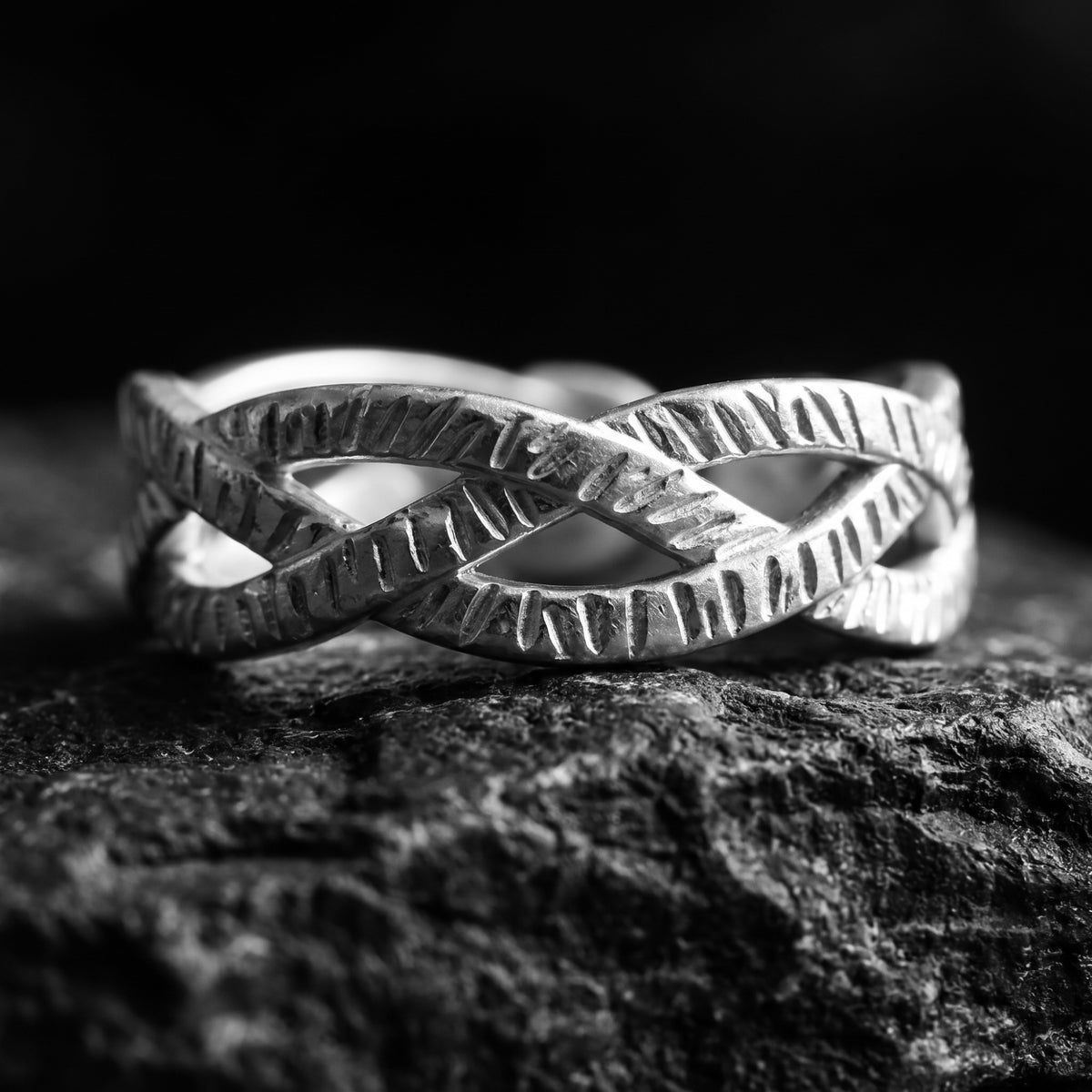 Close-up front view of a silver interlocking textured open ring resting on dark rock.