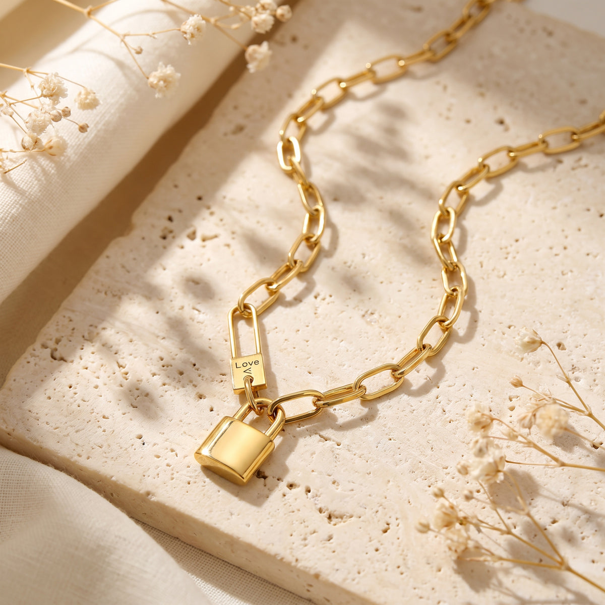 Gold-tone paperclip chain with a padlock pendant resting on a beige stone surface.