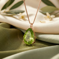 The Elyon Peridot Pendant Necklace in 18K Rose Gold, featuring a pear-cut natural peridot, resting on elegant green and cream silk fabric.