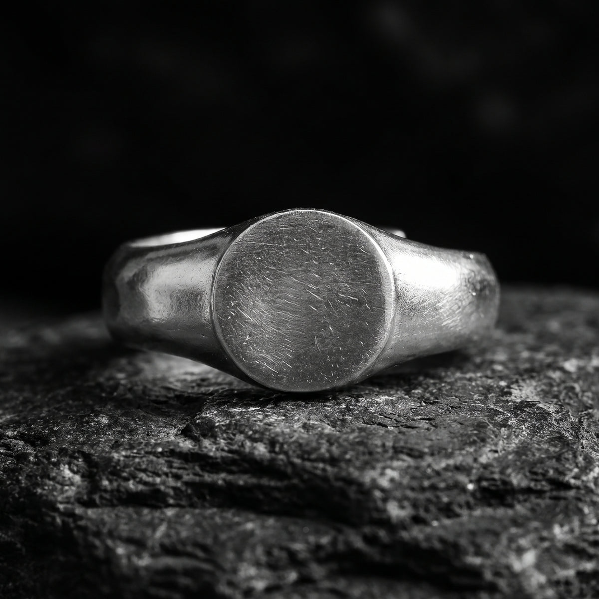 Close-up front view of a silver signet ring with a flat circular face on dark textured stone.