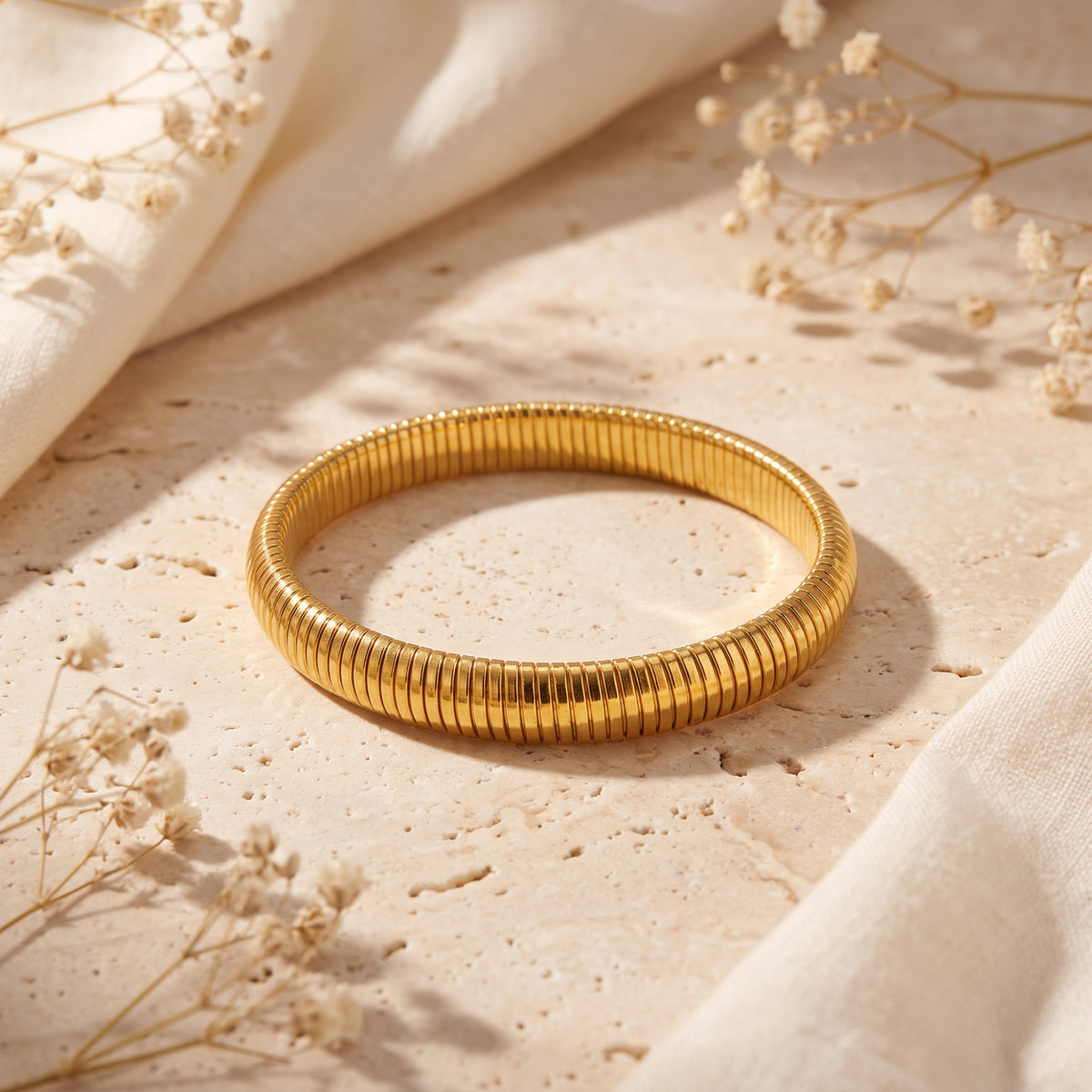 Gold-tone ribbed bracelet forming a closed loop on a travertine surface beside cream fabric and dried flowers.