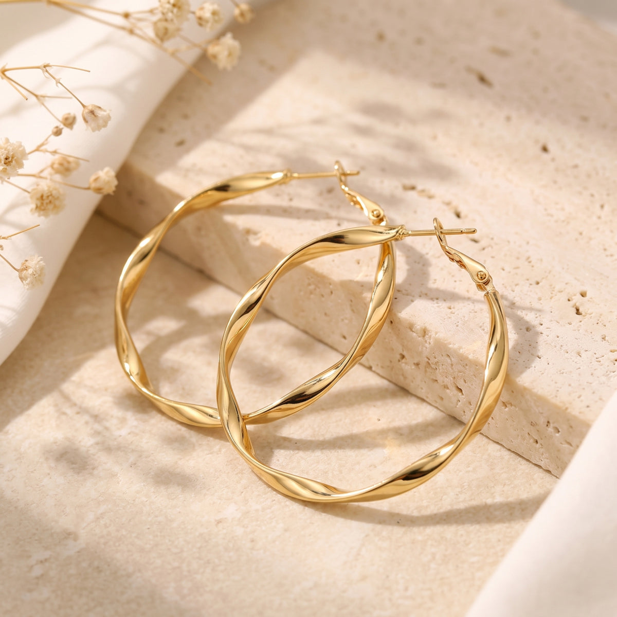 Pair of gold-finish twisted oval hoop earrings displayed on a textured stone surface with dried flowers.