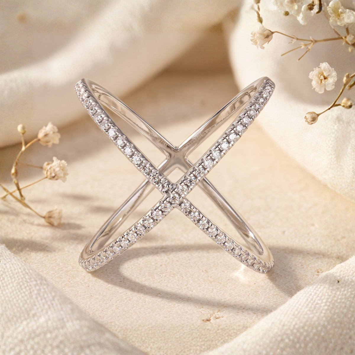 Silver X-shaped criss-cross ring adorned with clear pavé stones, standing upright in a soft neutral setting.
