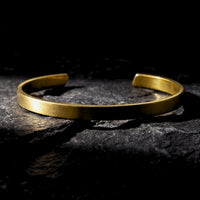 Gold stainless steel Meridian Cuff bracelet lying on a dark, textured rock surface.