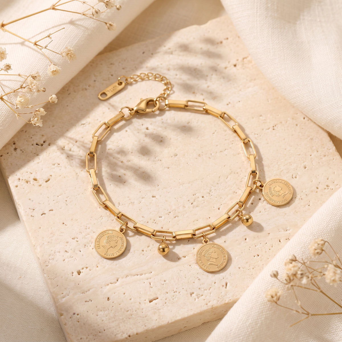 Gold-tone paperclip chain bracelet with coin and bead charms arranged on a textured stone slab.