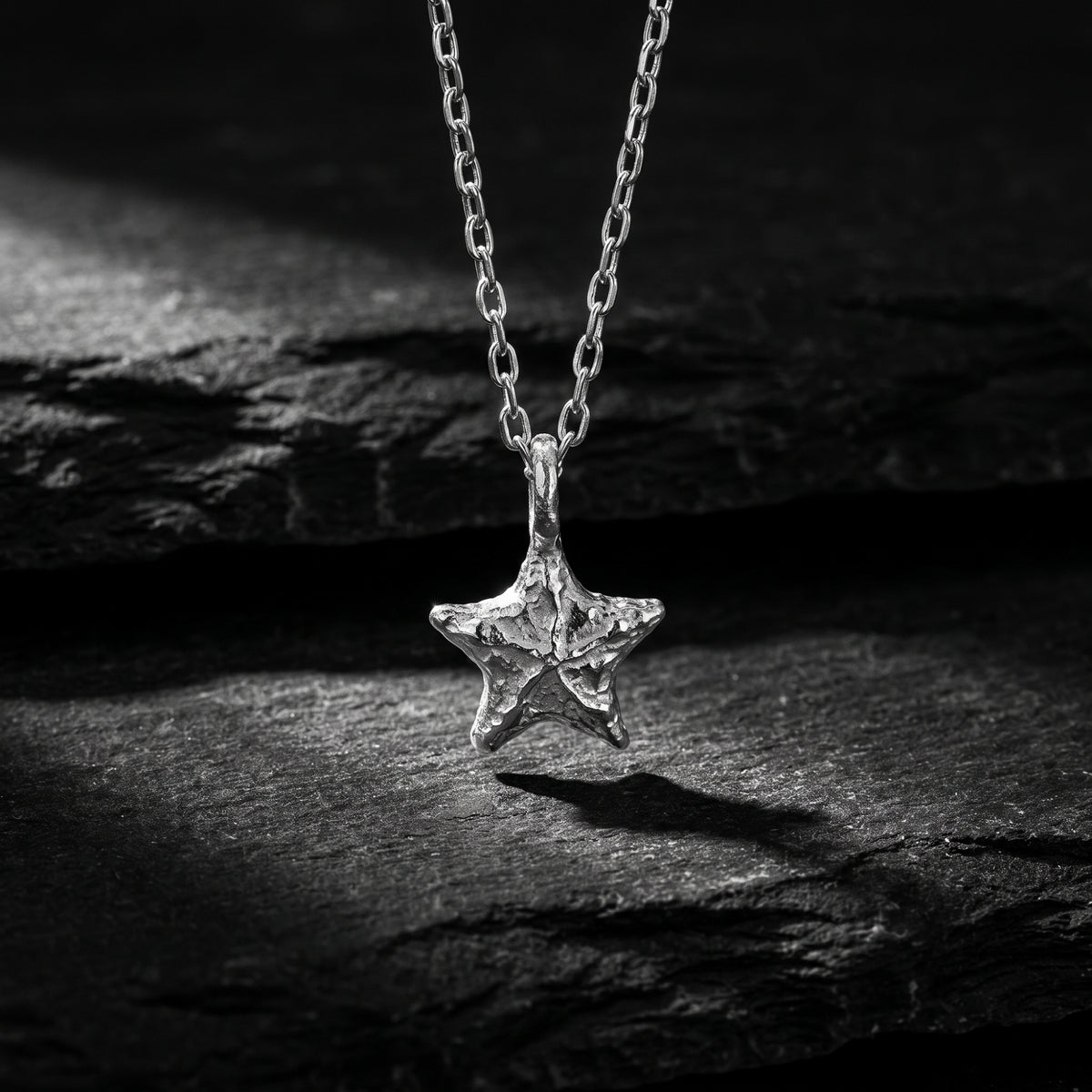 Textured silver starburst pendant suspended from its chain above a dark textured stone surface.