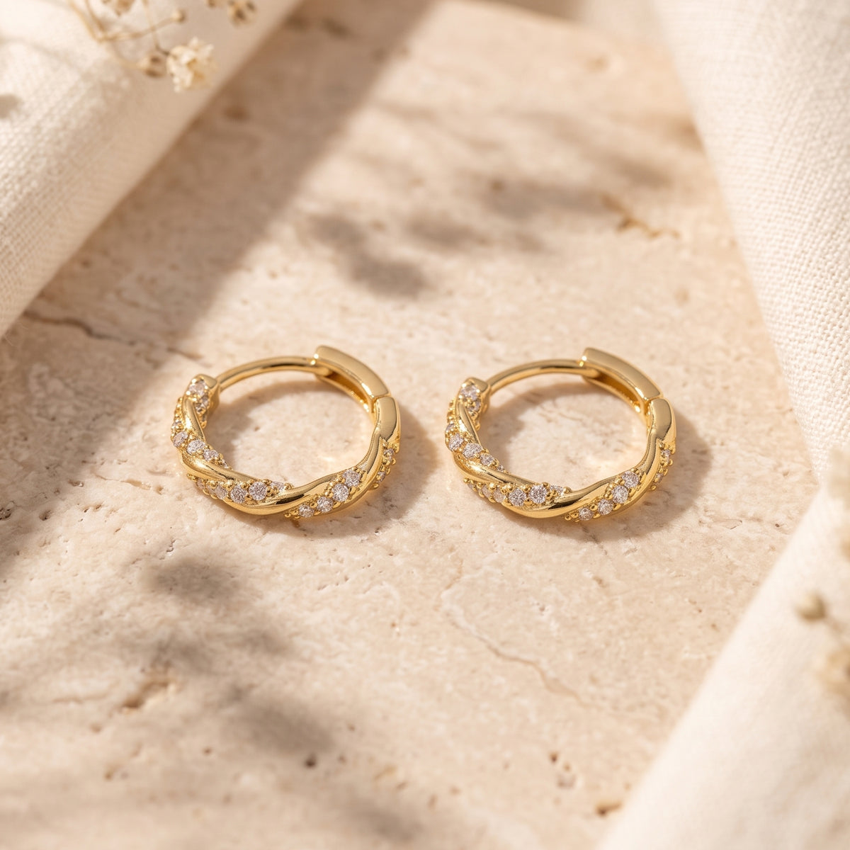 Gold-tone twisted huggie hoop earrings with pave-set clear cubic zirconia resting side by side on stone.