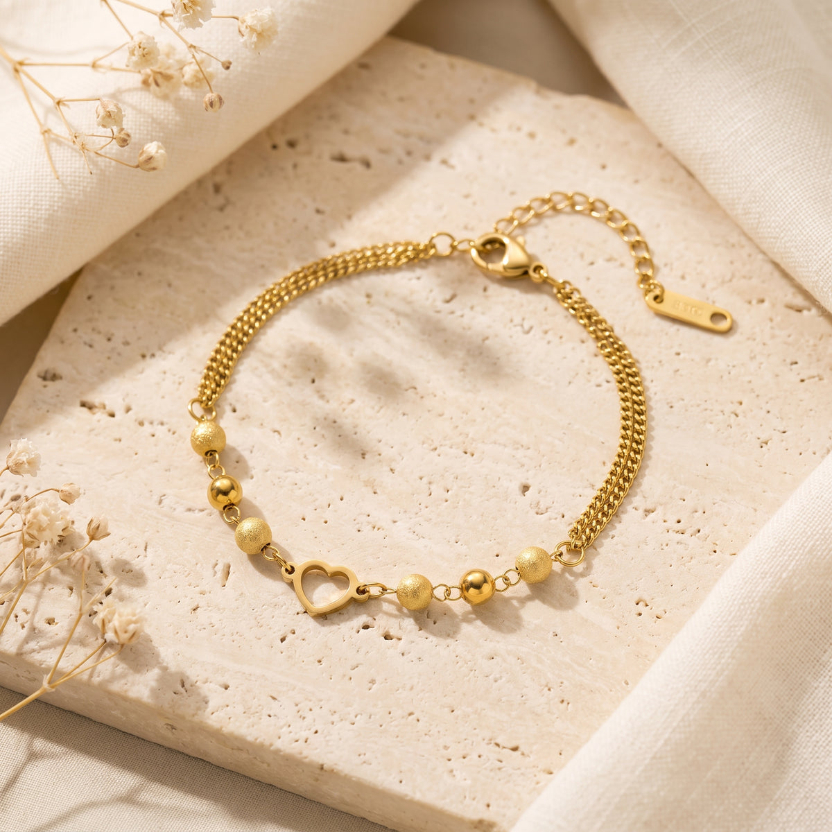 Gold-tone heart station bracelet laid flat on a beige stone slab with dried flowers, clasp and extender visible.