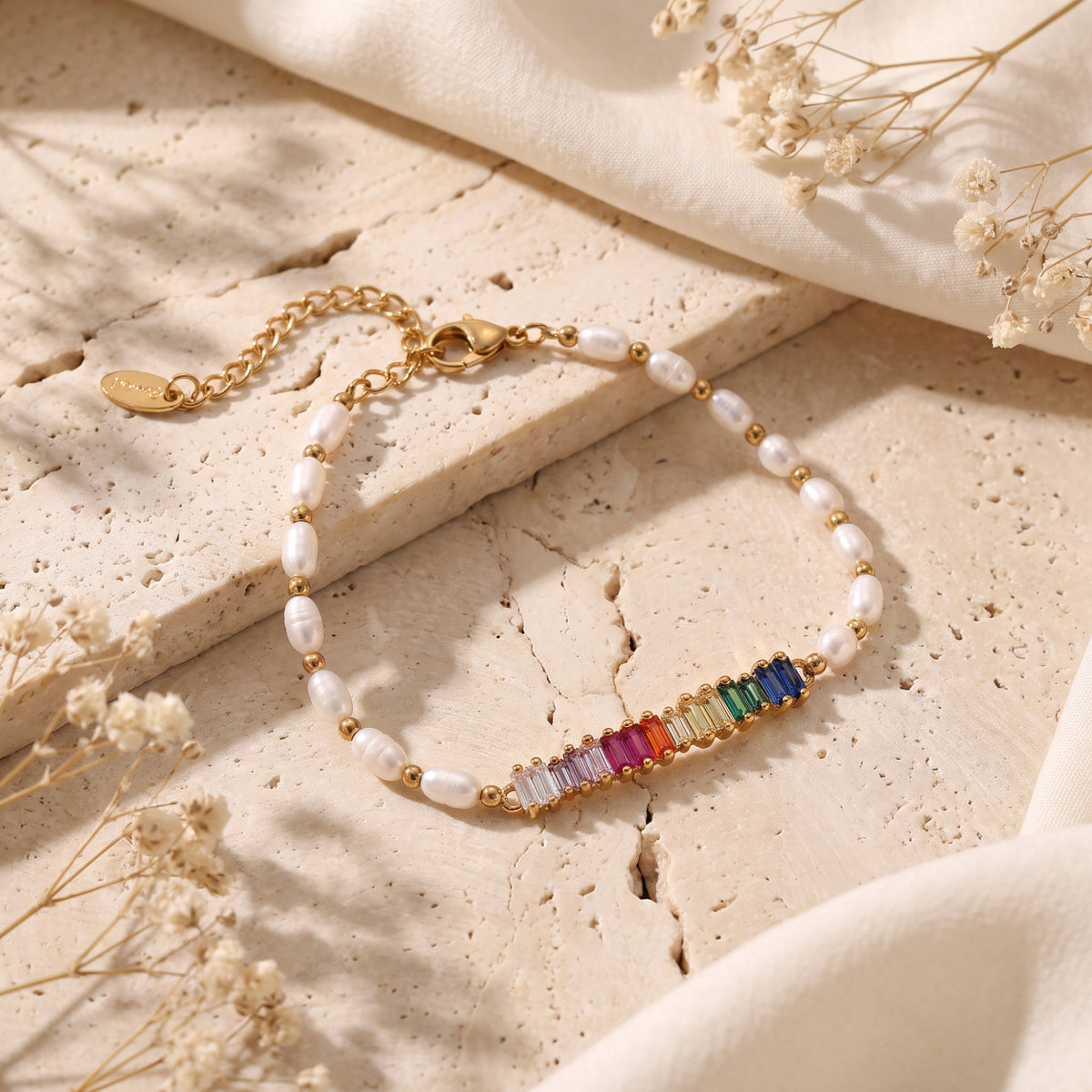 Gold-tone pearl bracelet with rainbow baguette stones laid flat on textured beige stone.