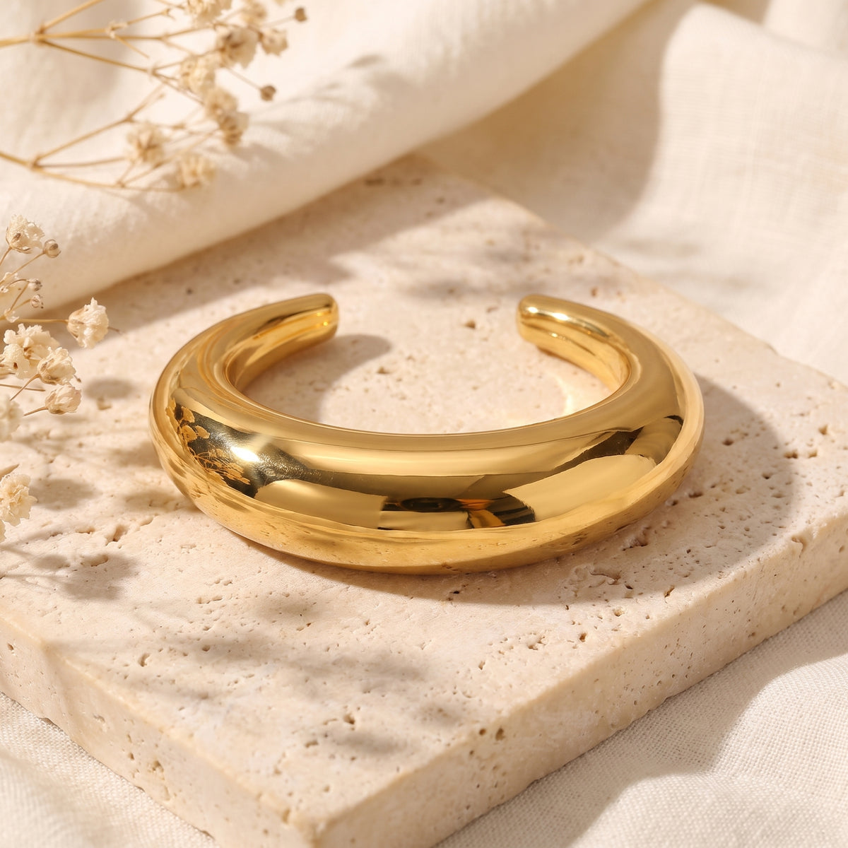 Smooth gold-toned open cuff bracelet with a thick dome shape displayed on a beige stone tile.