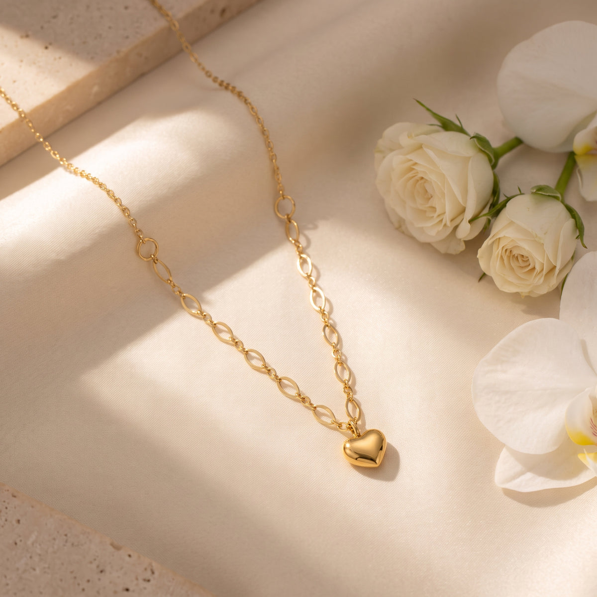 Gold-tone puffy heart necklace laid diagonally on cream fabric beside white roses and orchids.