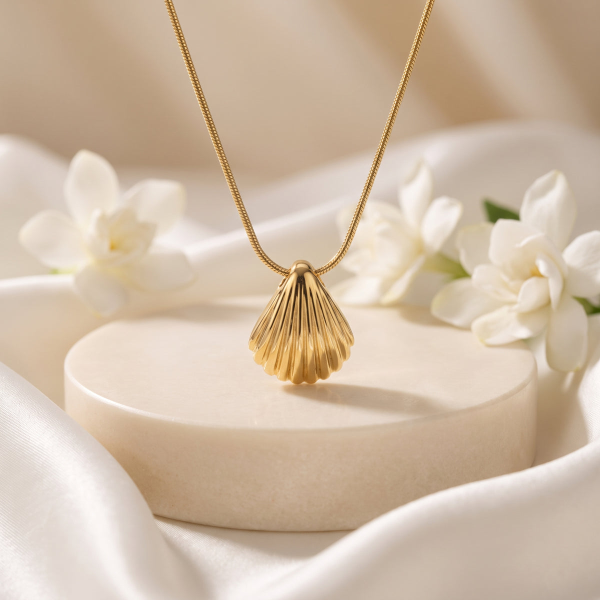 Gold scallop shell pendant necklace displayed above a round cream pedestal with white flowers and draped fabric.