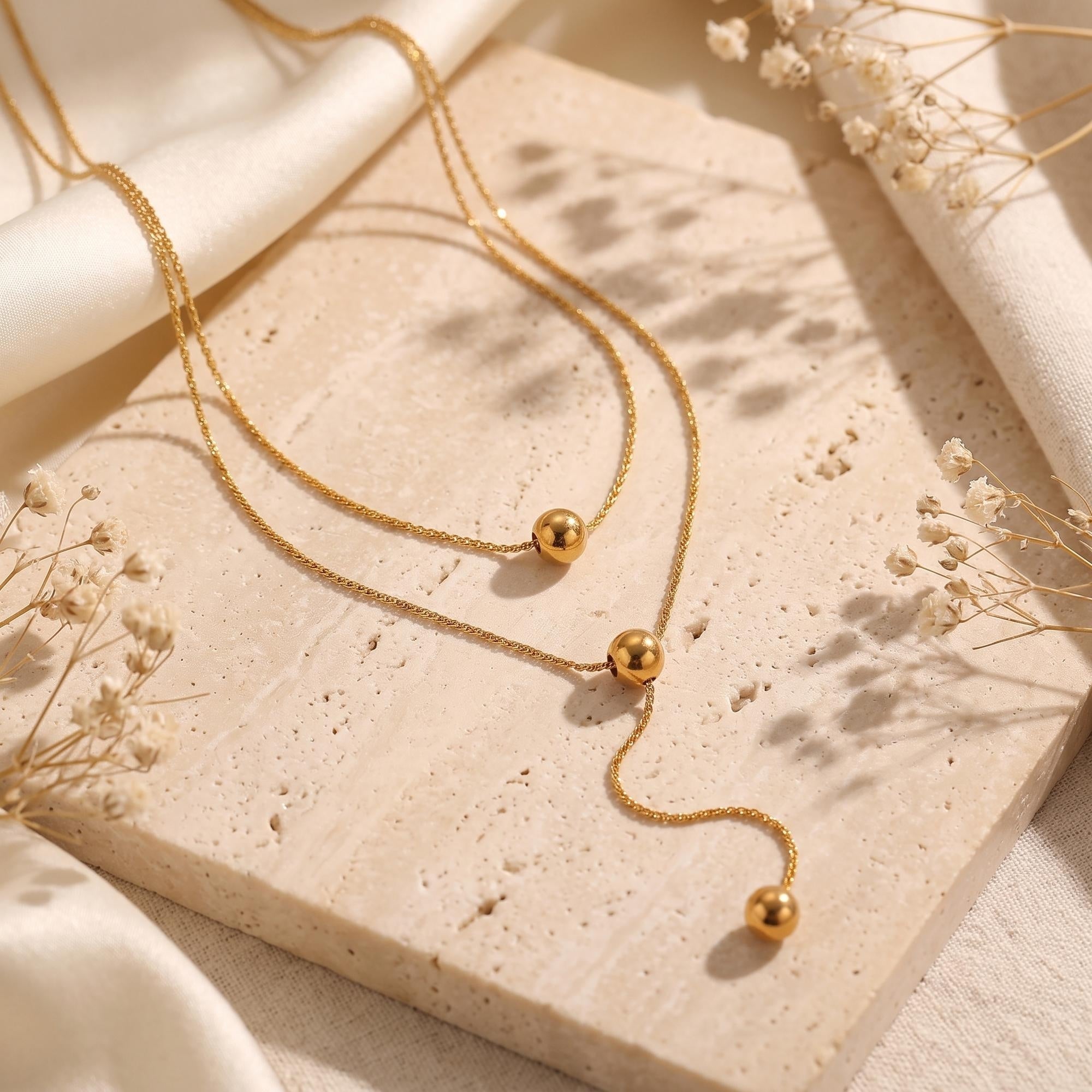 Gold layered lariat necklace with polished spheres displayed on a stone slab with dried flowers.