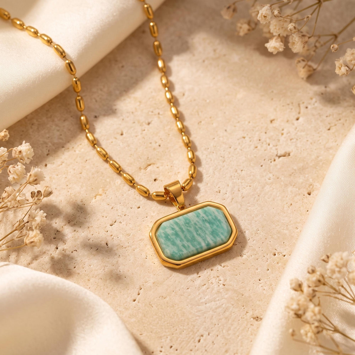Gold-tone bead chain necklace with a green Amazonite pendant on a beige textured surface with cream fabric and dried flowers.