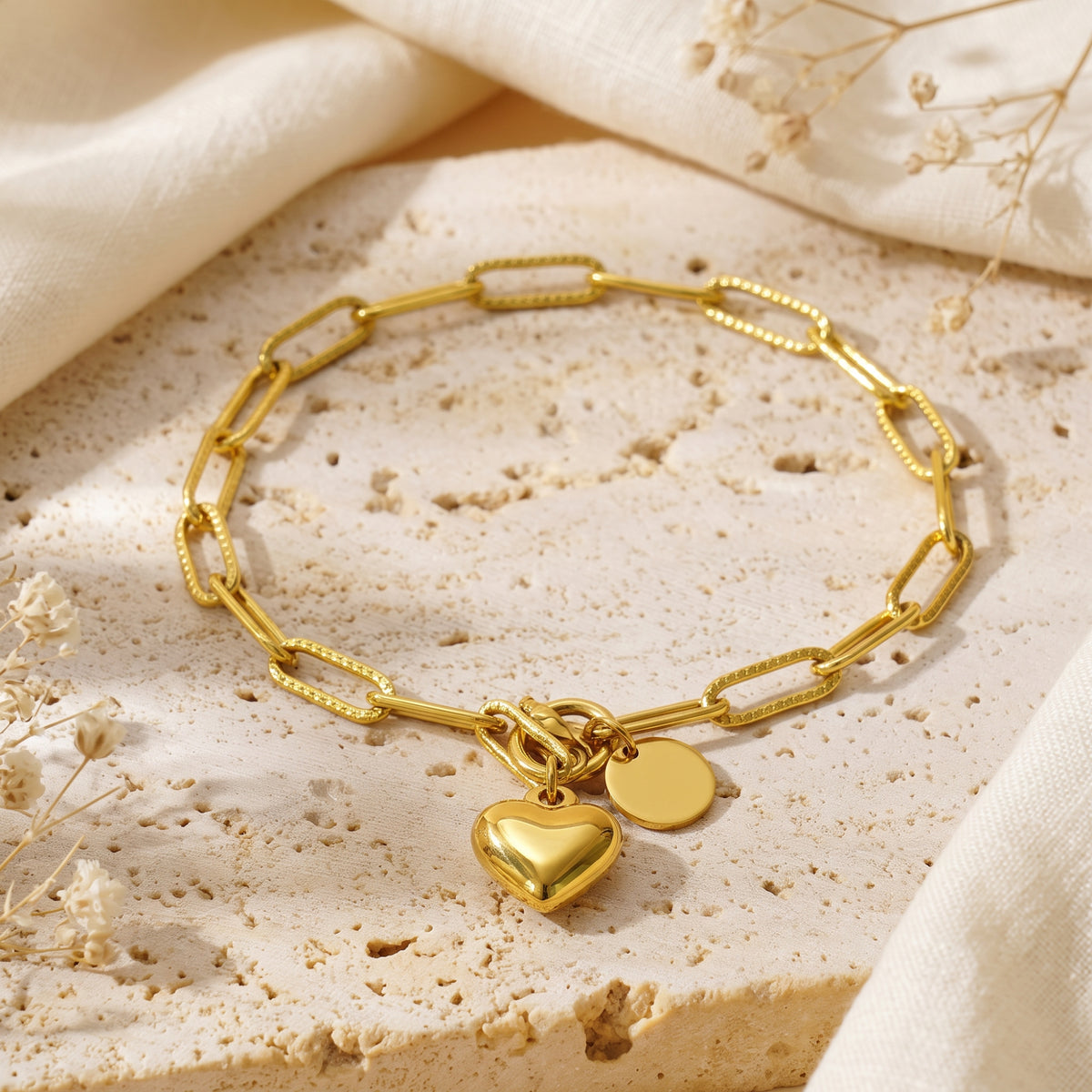 Gold-tone paperclip chain bracelet featuring a polished heart charm and flat disc charm laid on beige stone.