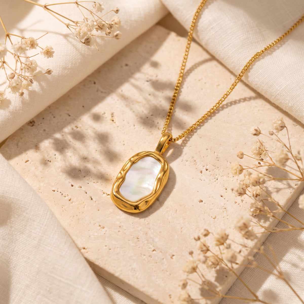 Gold-tone pendant necklace with a wavy bezel and shell inlay resting on a stone surface with dried flowers.