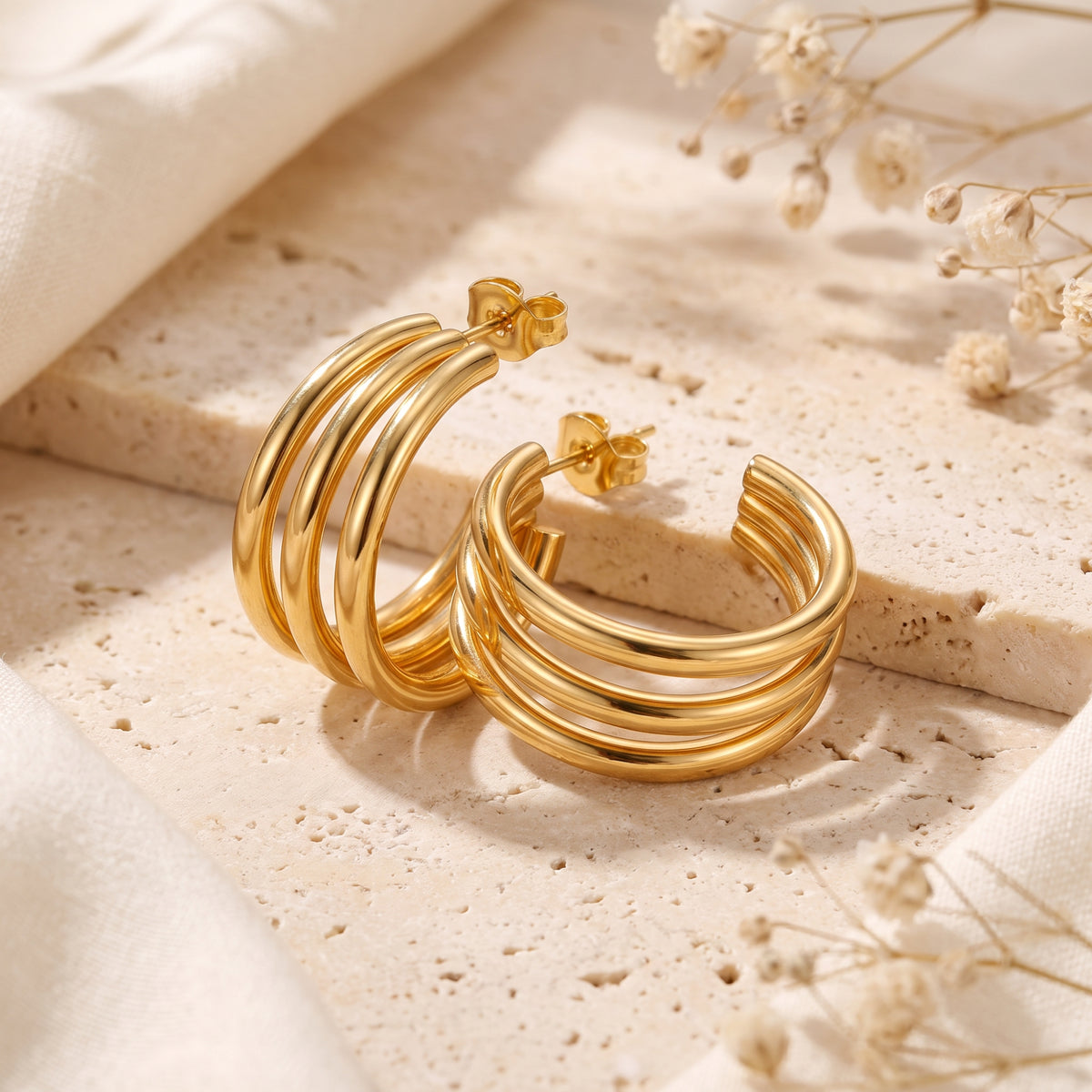 Gold-color triple-band C-hoop earrings with post backs displayed on a beige stone block alongside dried florals.