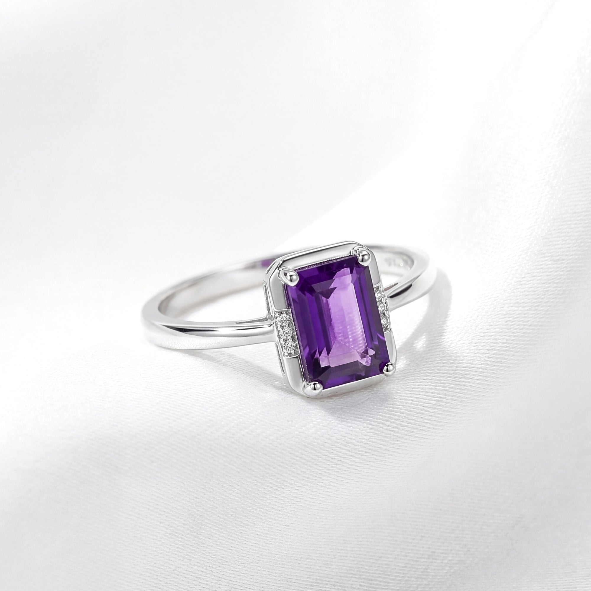 Emerald cut simulated amethyst solitaire ring in rhodium plated 925 sterling silver on white silk.