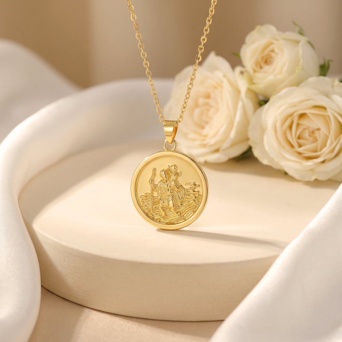 Gold-tone St. Christopher medallion necklace hanging from a beige display stand beside white roses.