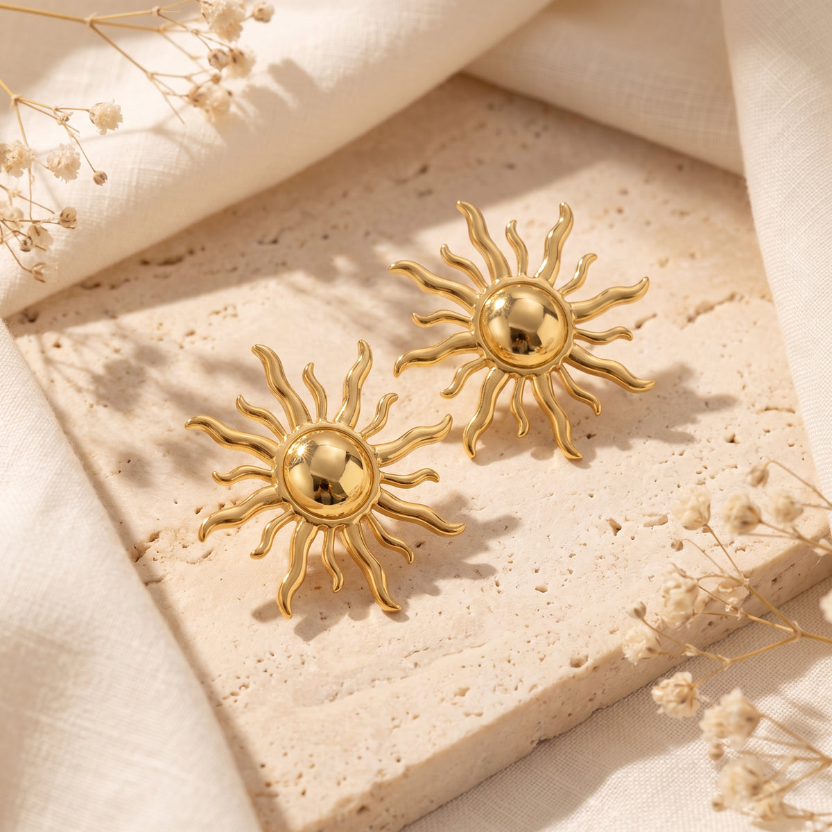Gold-tone sunburst stud earrings with wavy rays resting diagonally on a textured beige surface.