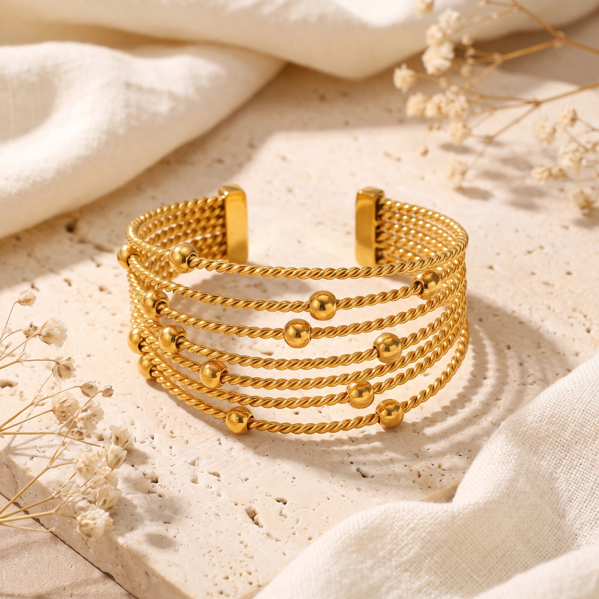 Gold-tone multi-strand twisted cable cuff bracelet with bead accents on a cream textured stone slab.