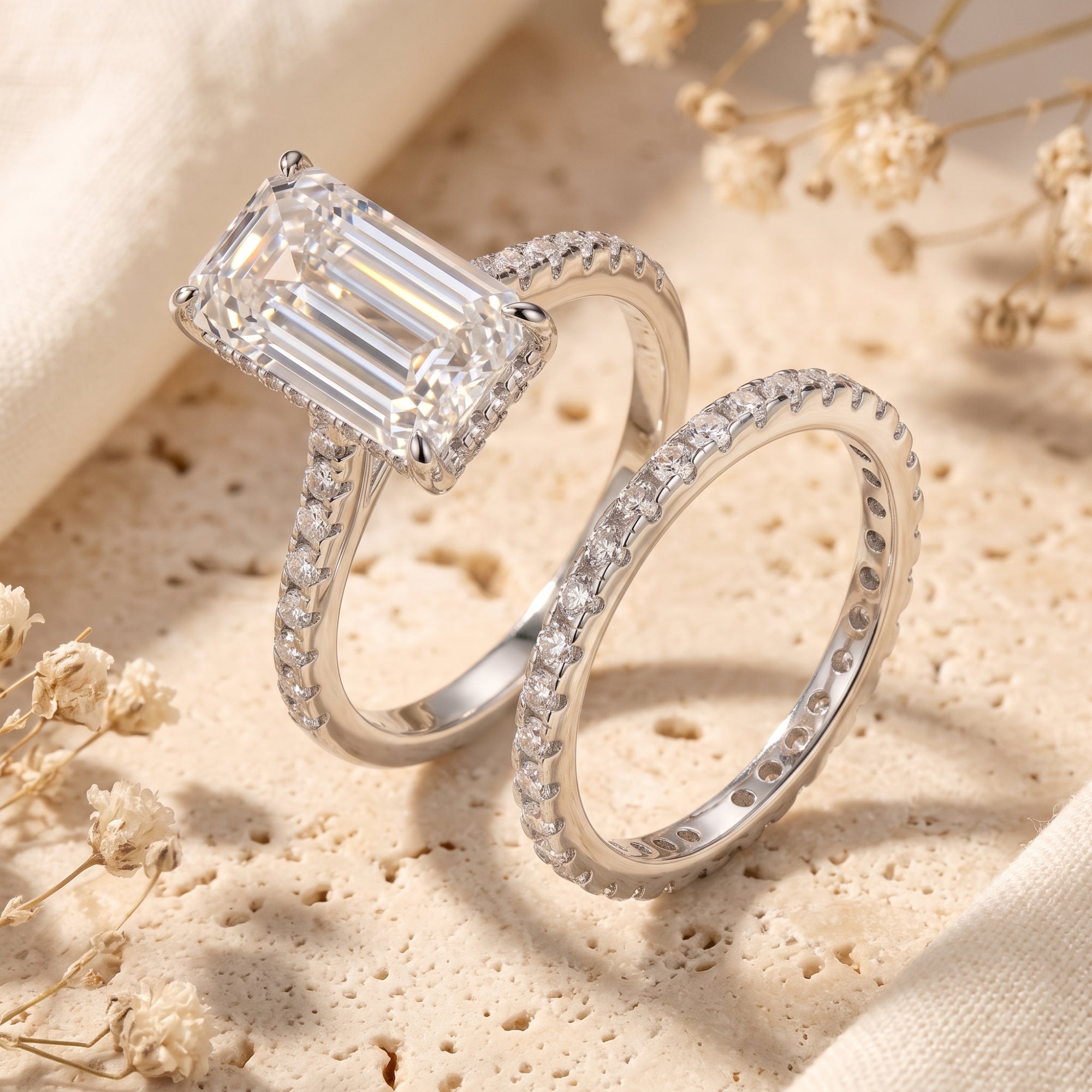 The Alaria emerald-cut simulated diamond ring and matching silver band on a stone surface with dried flowers.