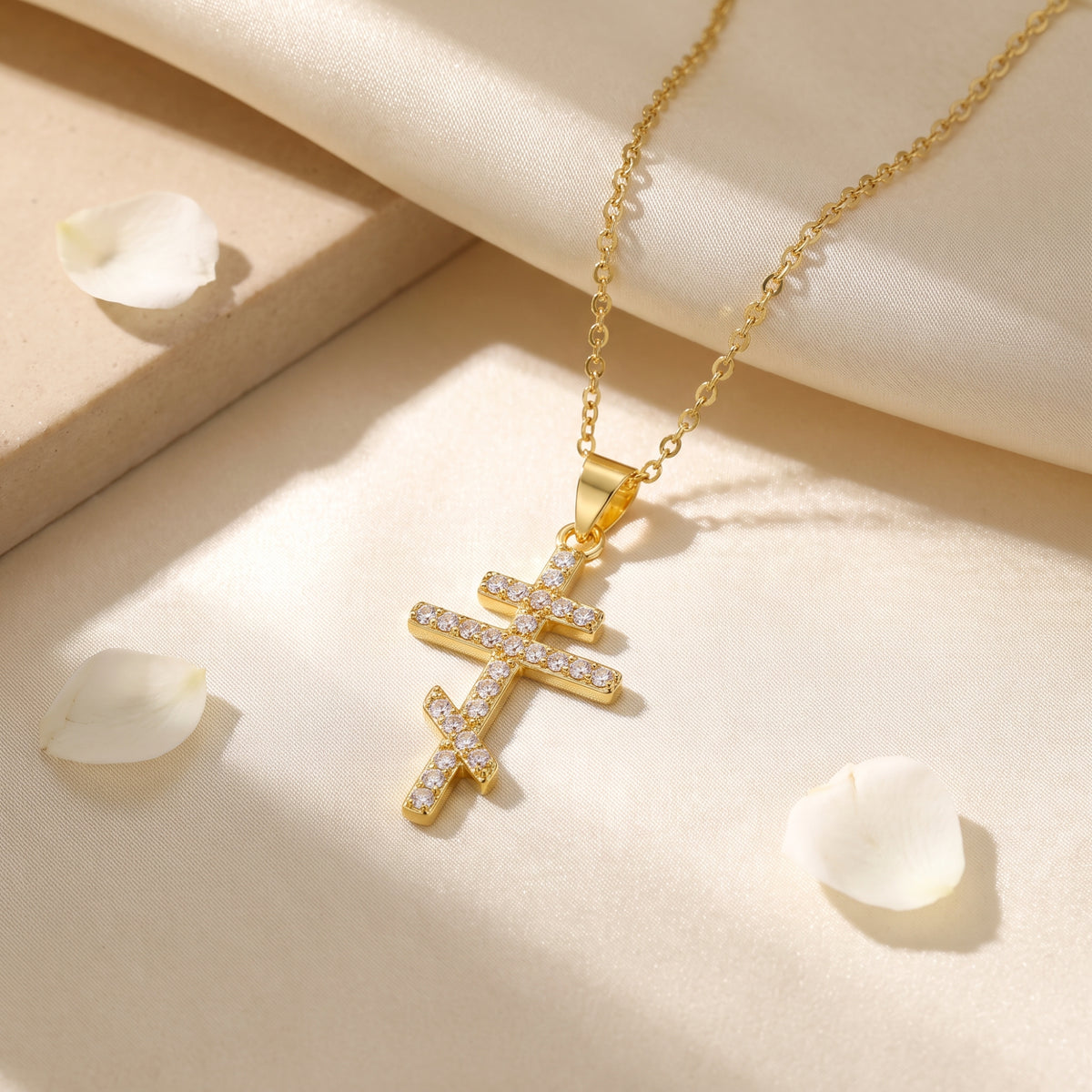 Gold-tone Orthodox cross pendant with clear stones on cream silk, surrounded by white petals.