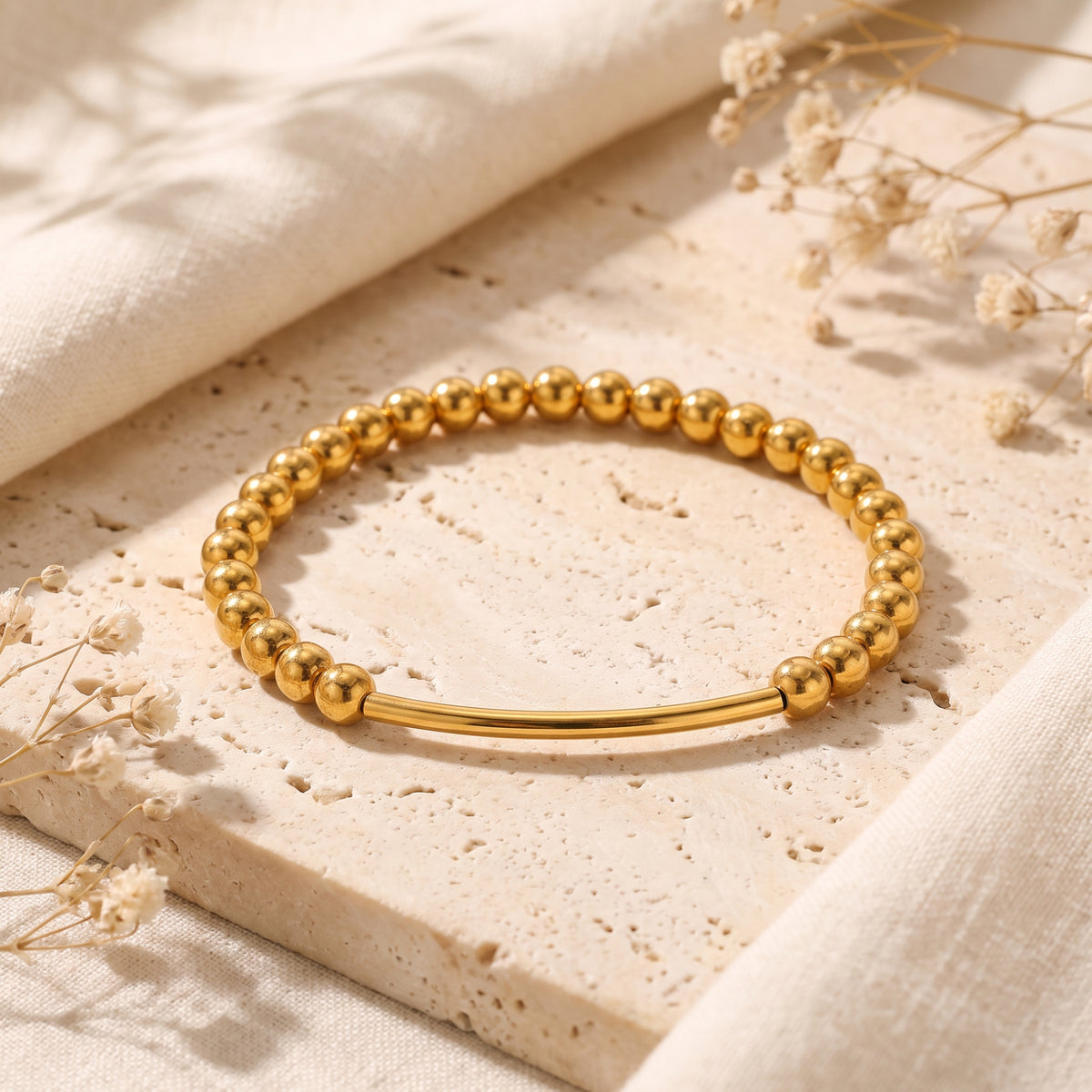 Gold-tone beaded bracelet with a curved bar centerpiece on a textured beige stone slab.
