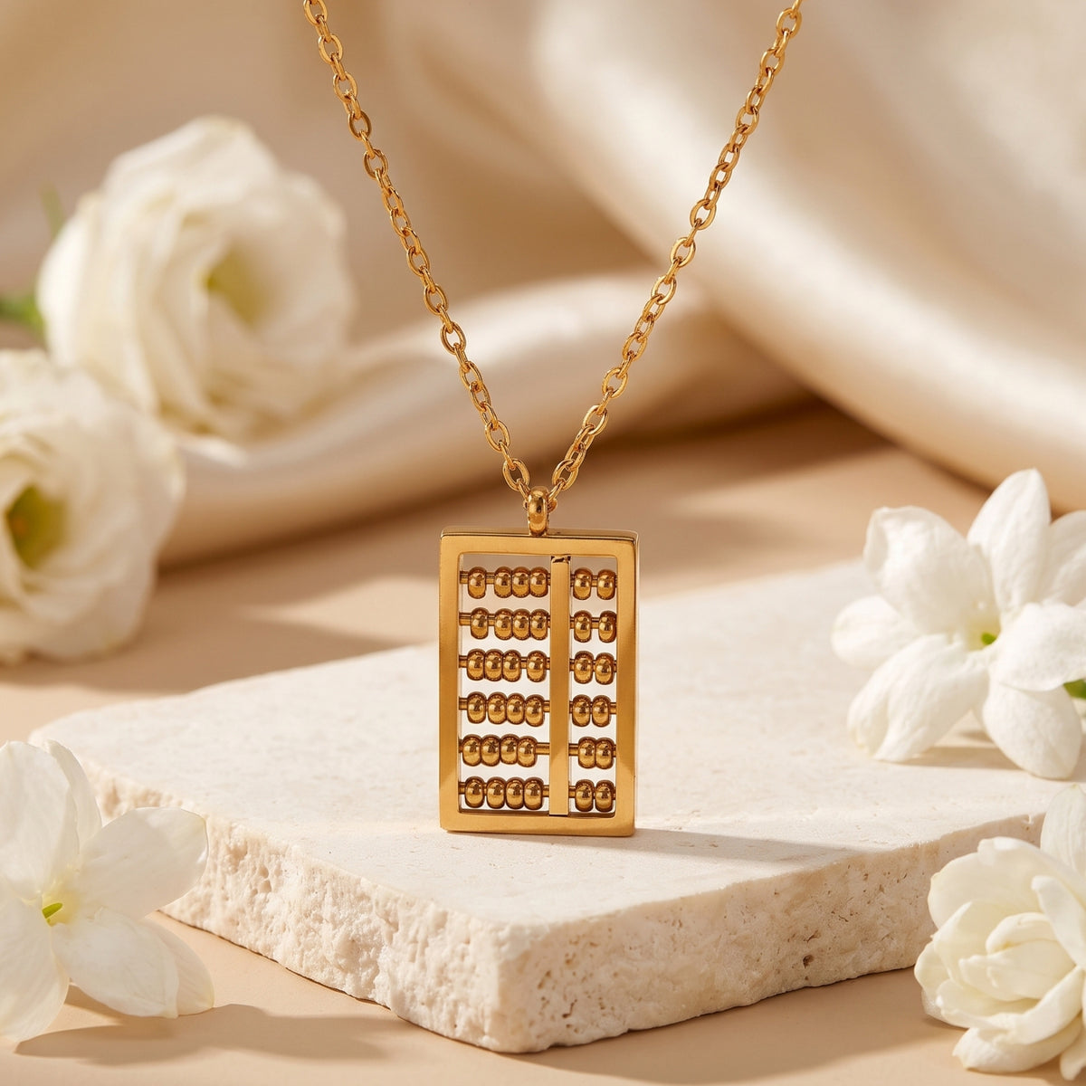 Gold-tone rectangular abacus pendant necklace displayed upright on a beige stone block with white flowers.