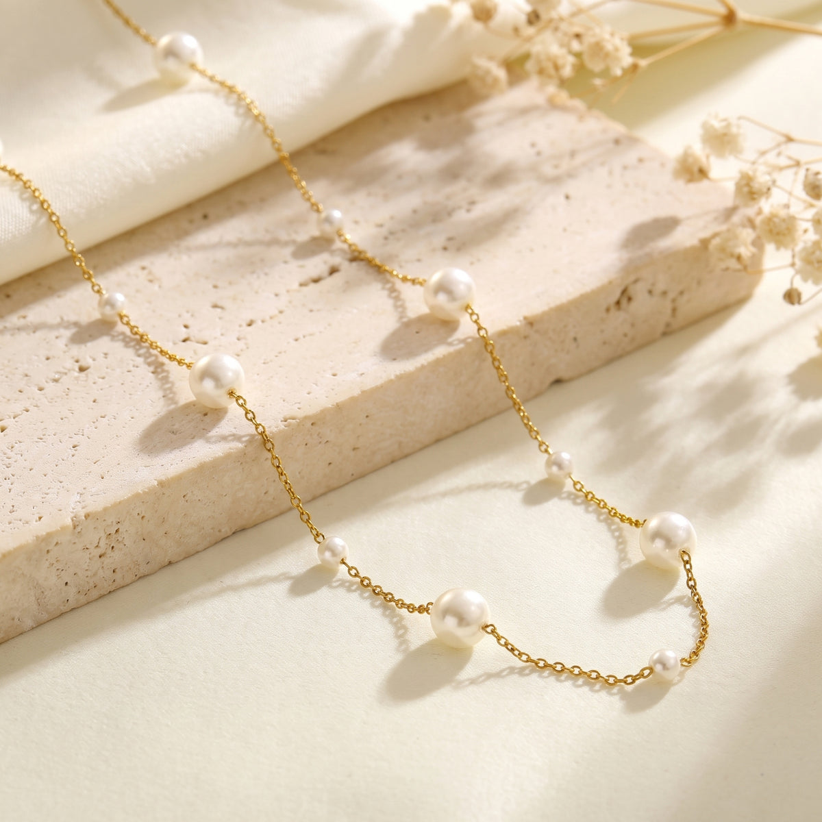 Gold-tone pearl station necklace arranged on a textured stone slab beside cream fabric.