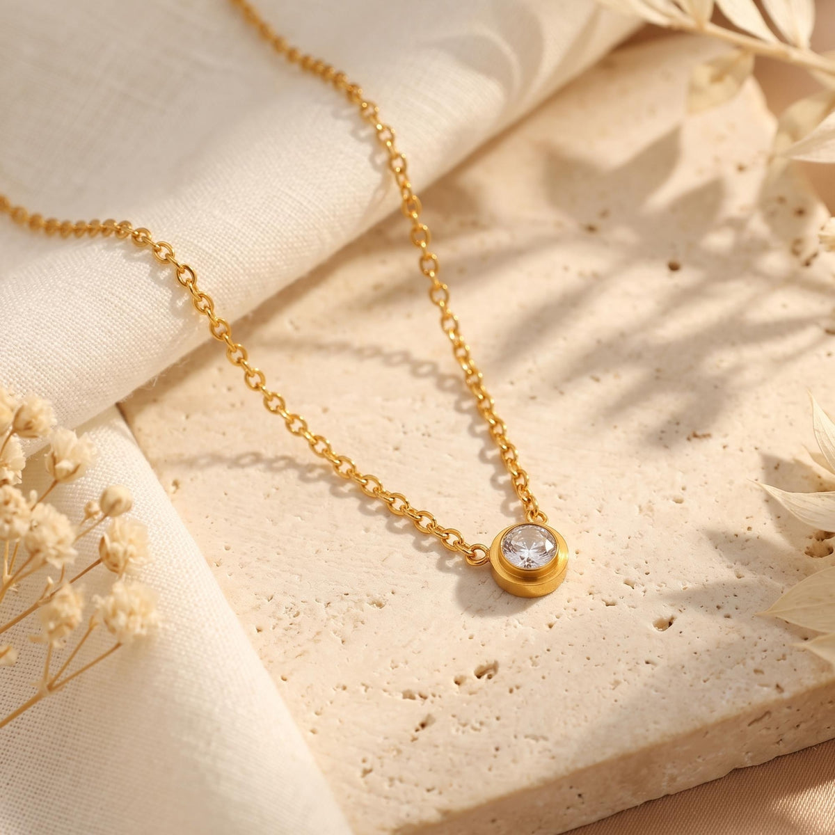 Gold-tone bezel solitaire necklace with a clear round center stone on a pale stone surface beside white fabric.