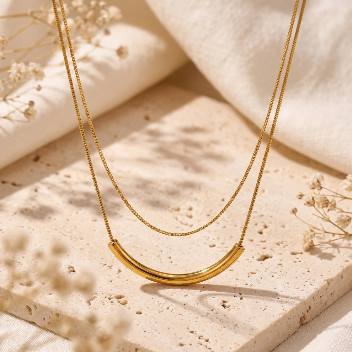 Gold-tone layered necklace with a curved arc bar pendant suspended over textured stone and dried flowers.