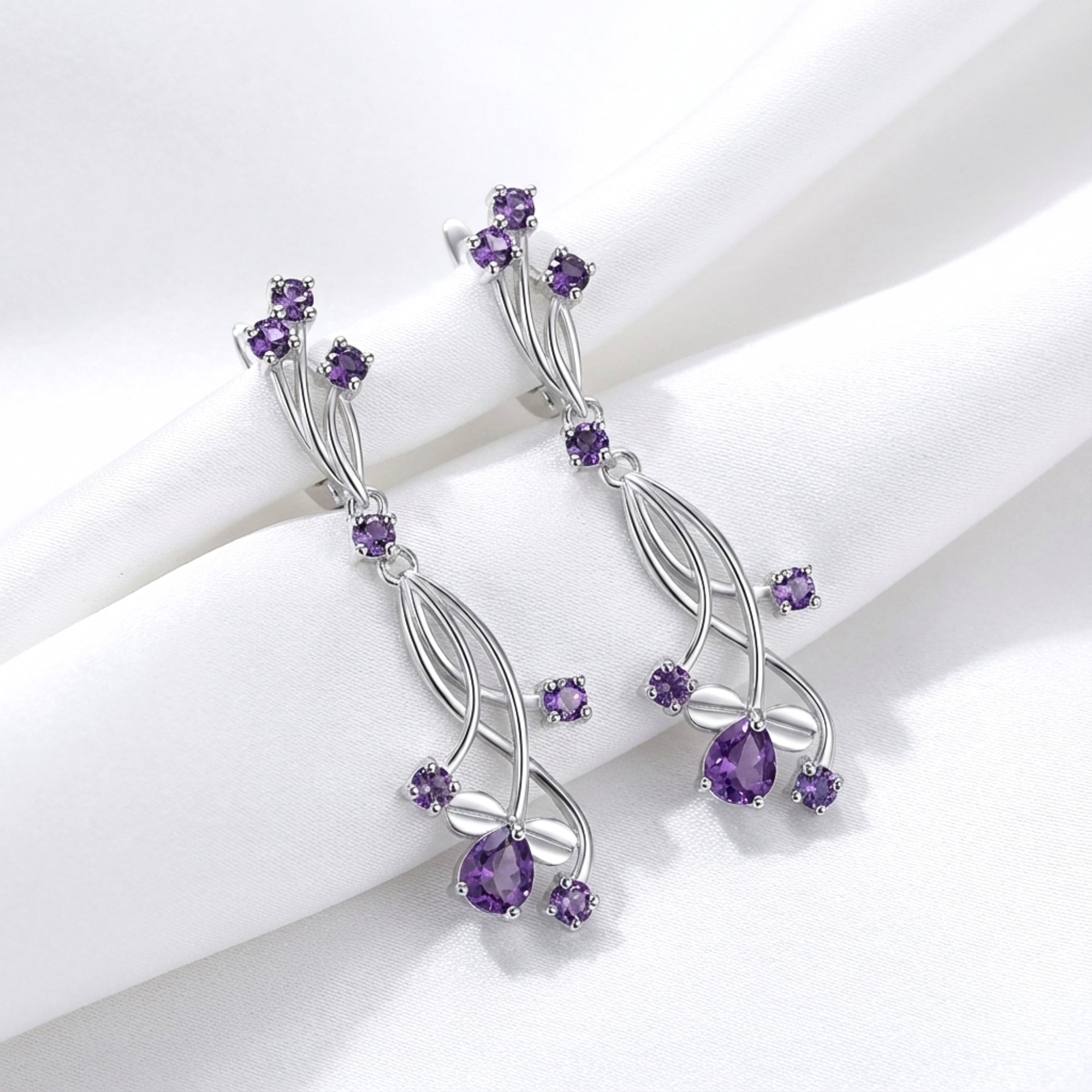 Lyriel drop earrings in S925 sterling silver with pear and round-cut simulated amethyst on white silk fabric.