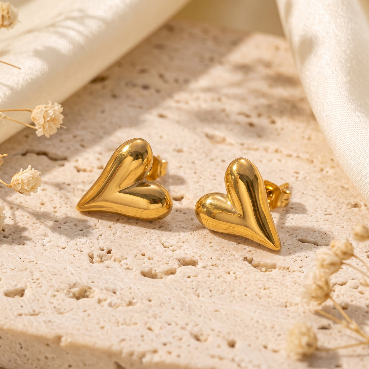 Gold-colored puffed heart stud earrings rest on a textured stone surface with delicate white flowers.