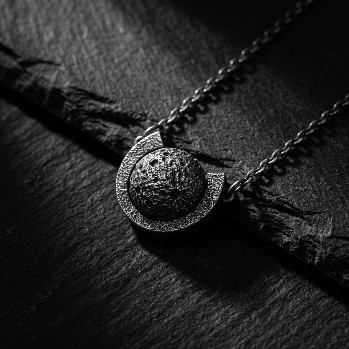 Angled macro shot of the textured silver lunar sphere and arc pendant resting on dark slate.