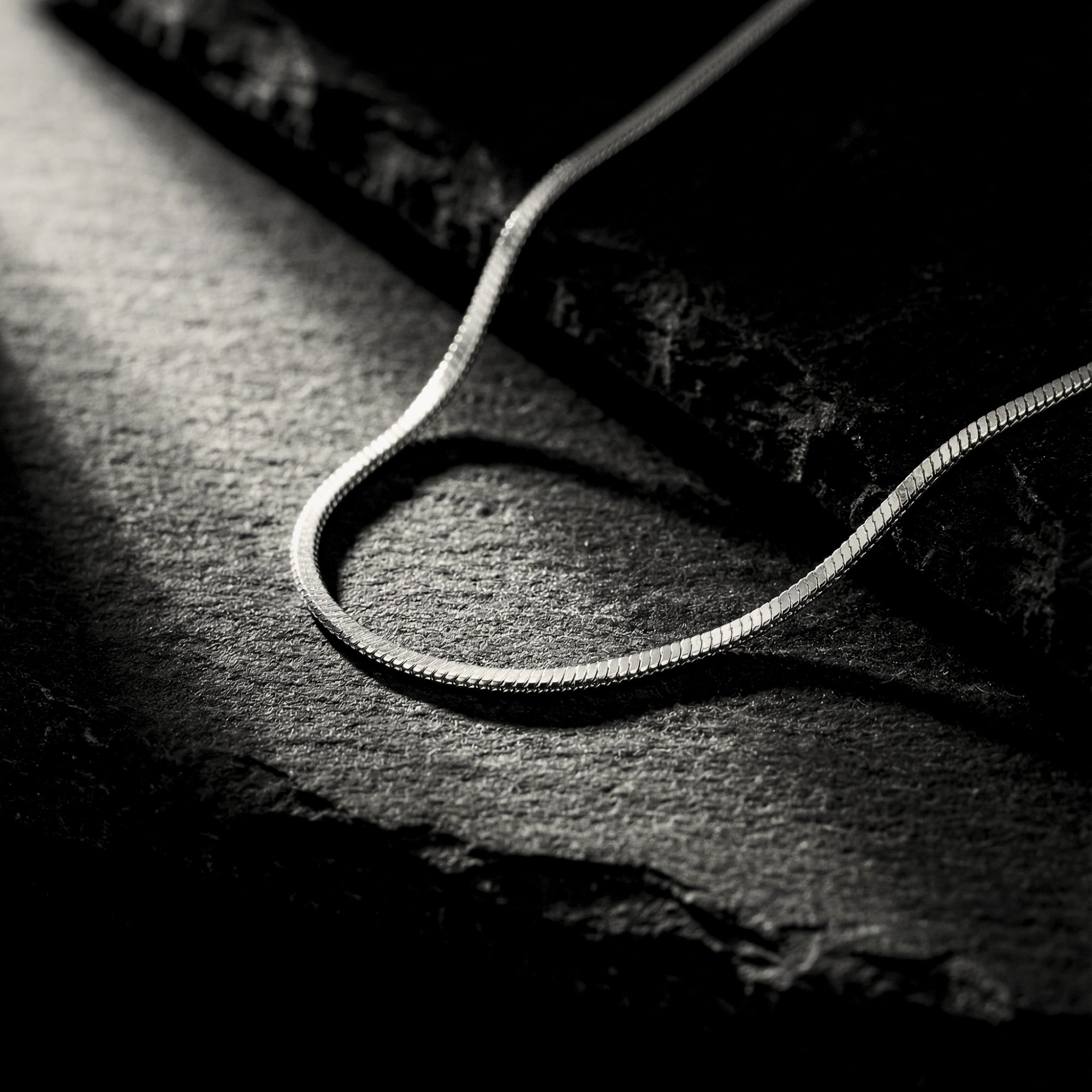 Silver stainless steel square link chain necklace draped on a dark, textured stone surface with dramatic lighting.