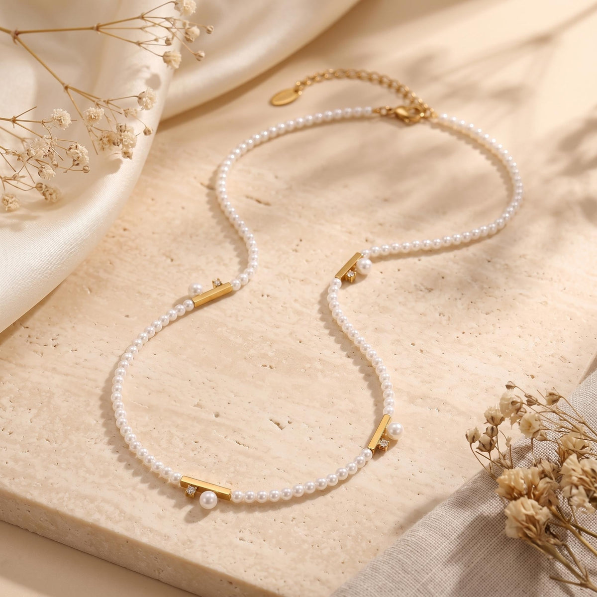 Flat lay of the gold-tone pearl bar station necklace on a travertine surface with dried flowers and cream fabric.