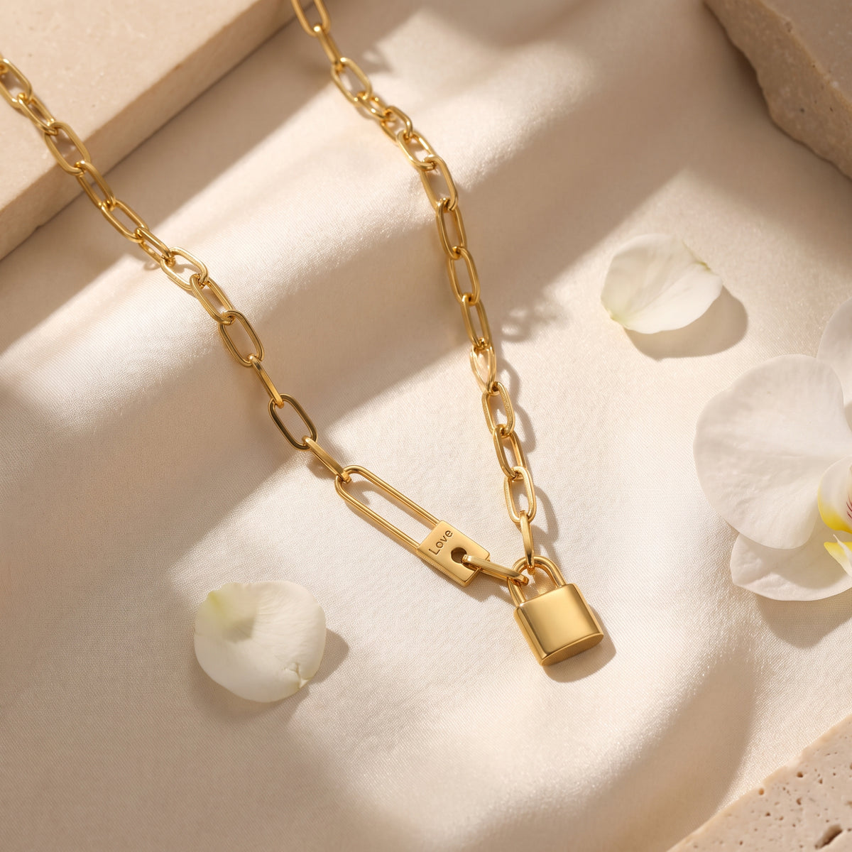 Gold-tone paperclip chain necklace with padlock pendant and “Love” engraving, laid on fabric among flower petals.