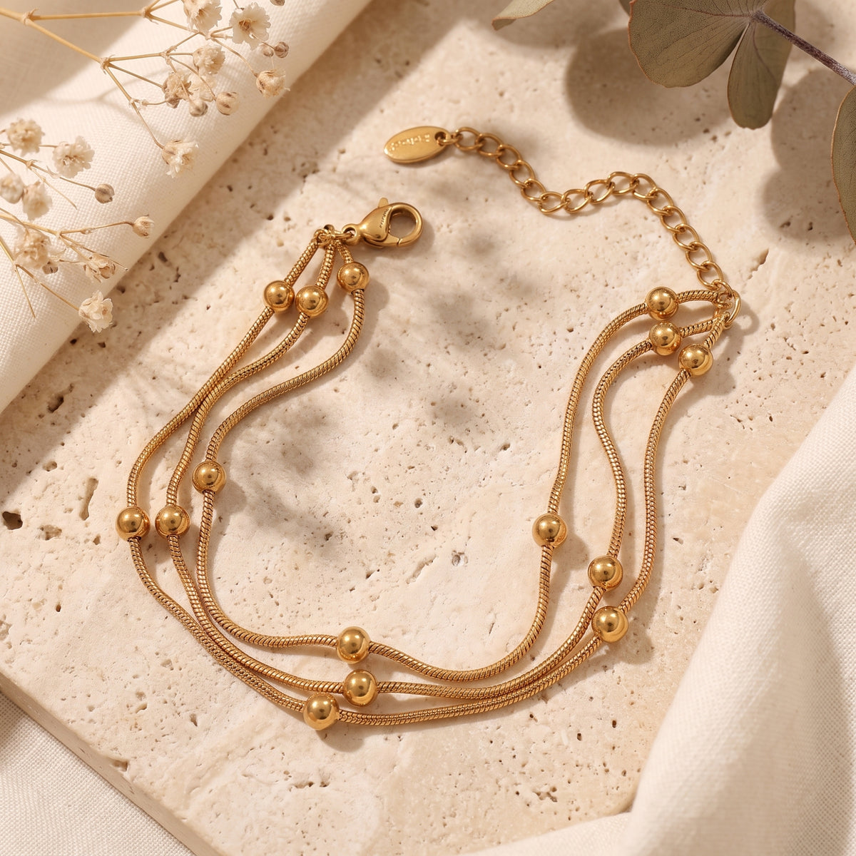 Gold three-strand snake chain bracelet with polished sphere beads, laid on stone beside dried flowers.