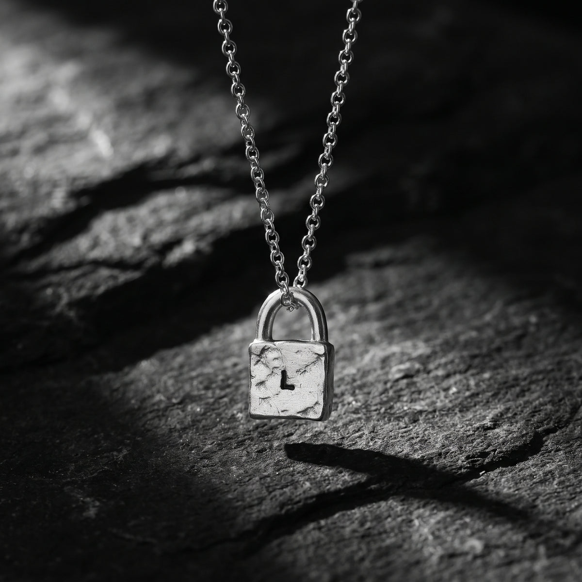 Front view of a textured silver lock pendant stamped with the letter L, hanging above dark slate.