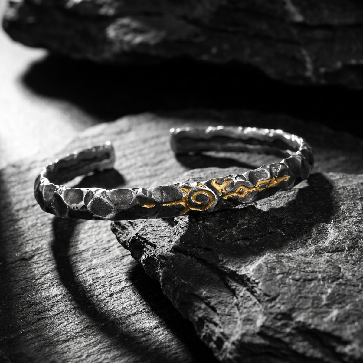 Hammered silver cuff bracelet with a central gold-tone sigil, resting on dark slate stone.
