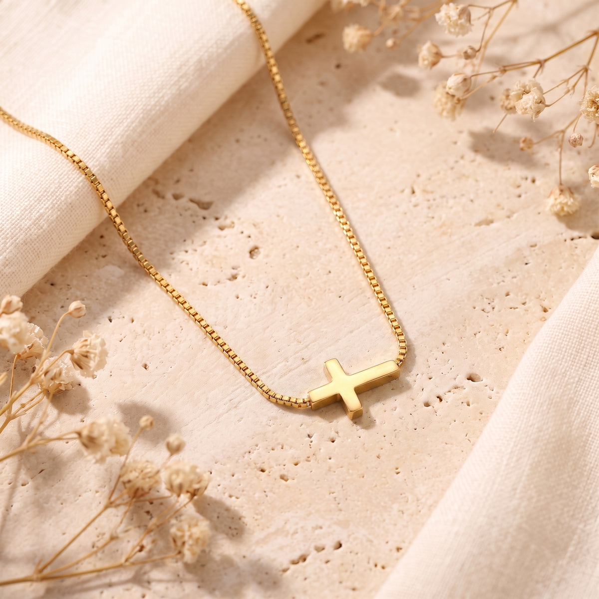 Gold-tone sideways cross pendant on a box chain lying flat on textured stone beside white fabric.