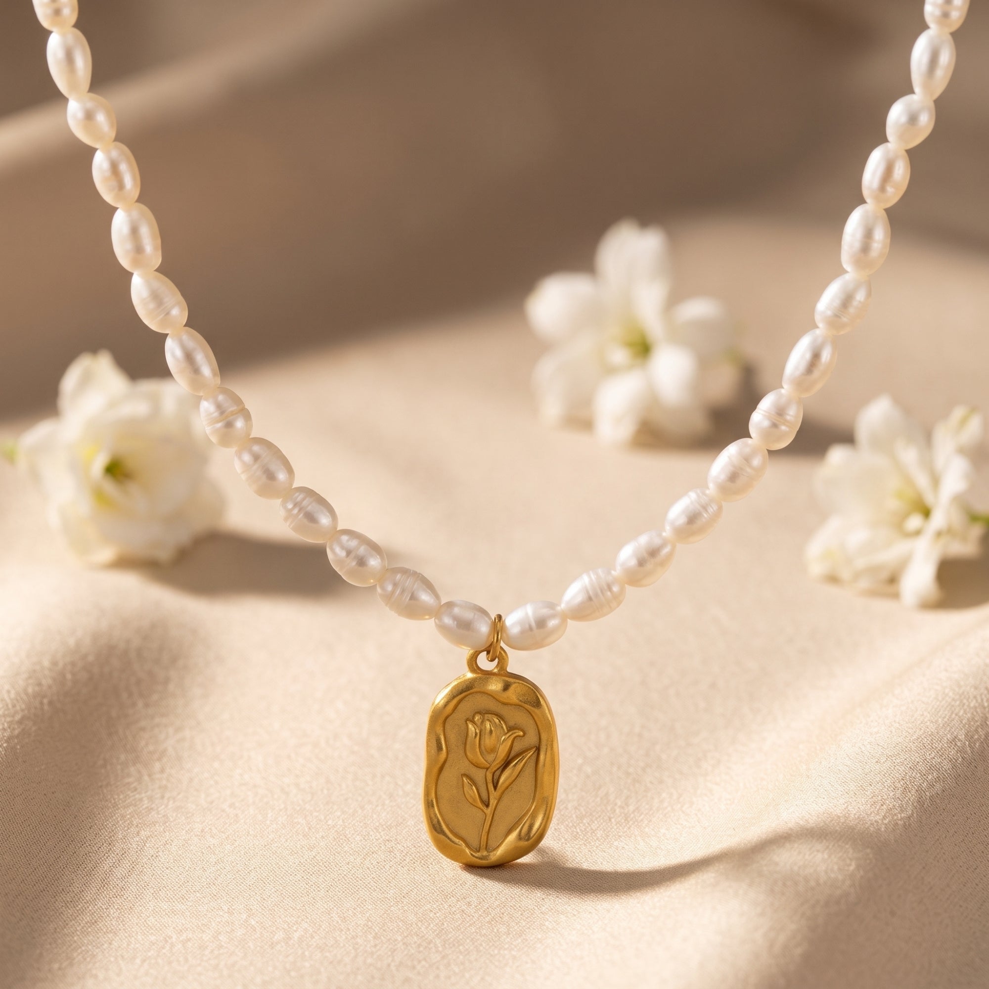 Rosalinde pearl necklace with a gold rose pendant on a soft fabric background with white flowers.