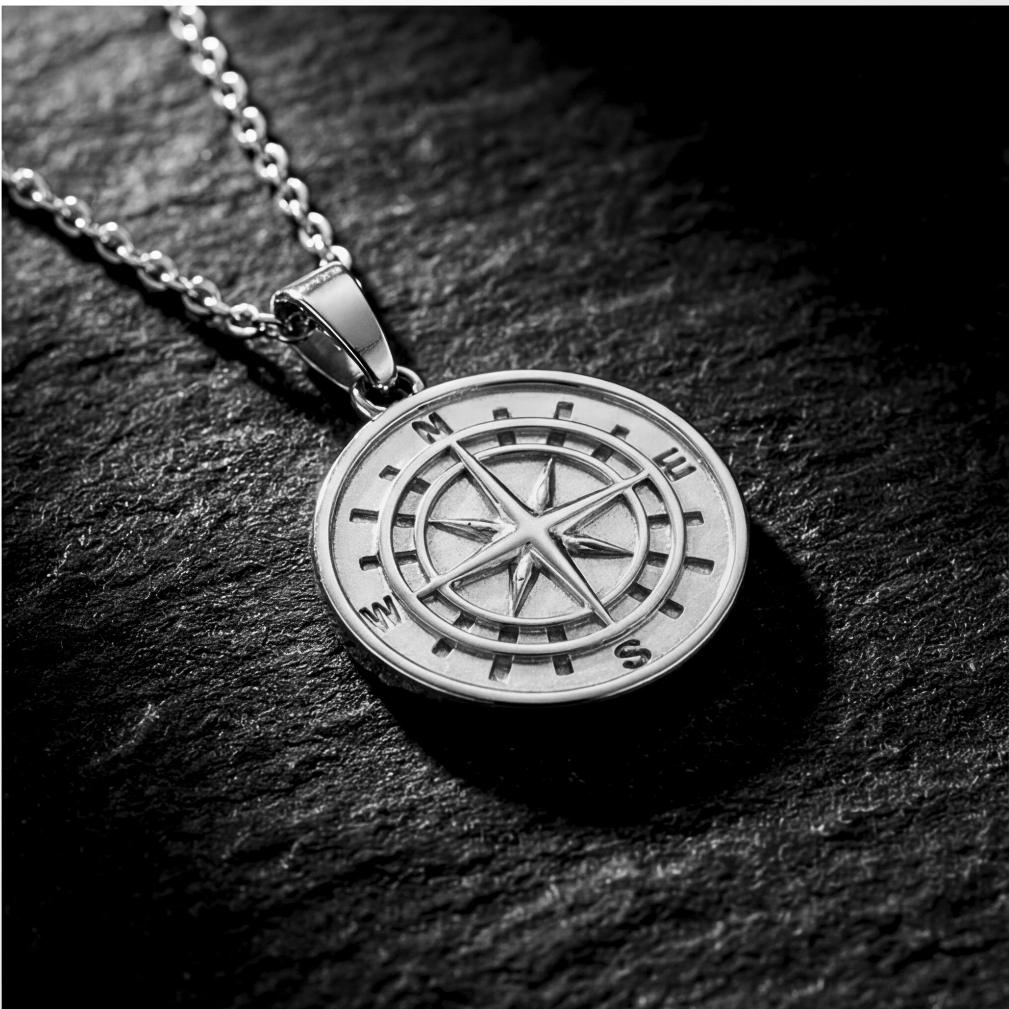 Silver stainless steel compass pendant on a chain, resting on a dark textured stone surface.
