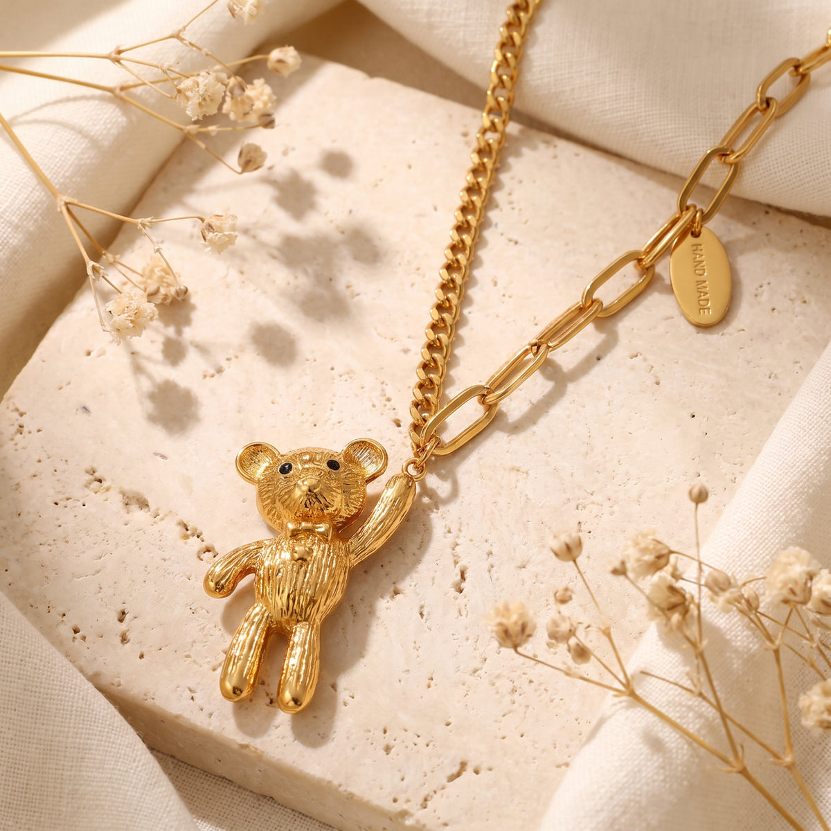 Close-up of a textured gold-tone teddy bear pendant resting on a mixed-link chain atop a stone slab.