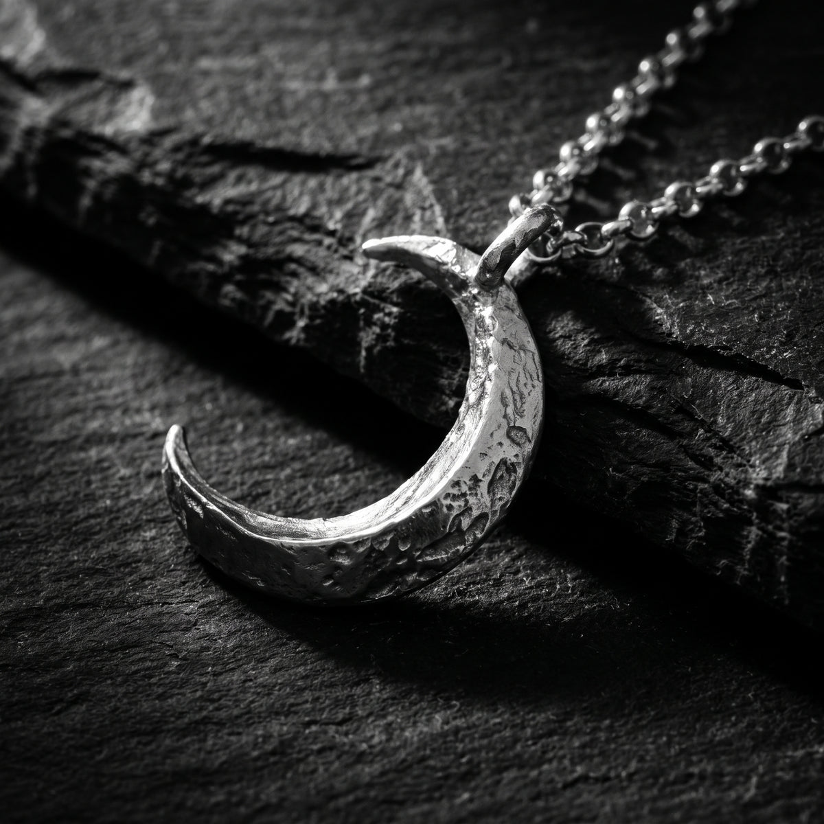 Close-up of a silver textured crescent moon pendant lying on dark slate stone.