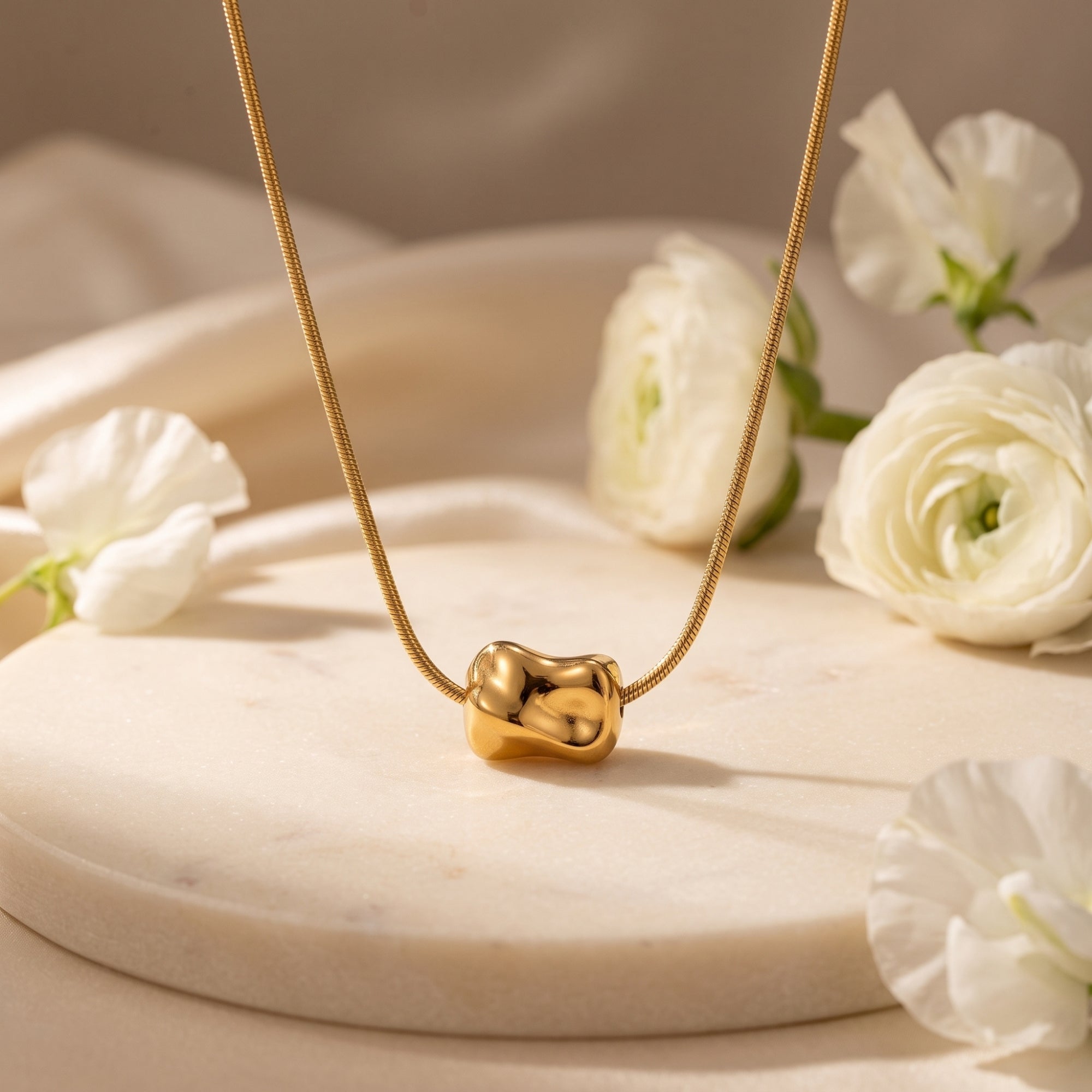 18K gold finish Petra necklace with a molten sculptural bead on a snake chain, styled with white flowers.