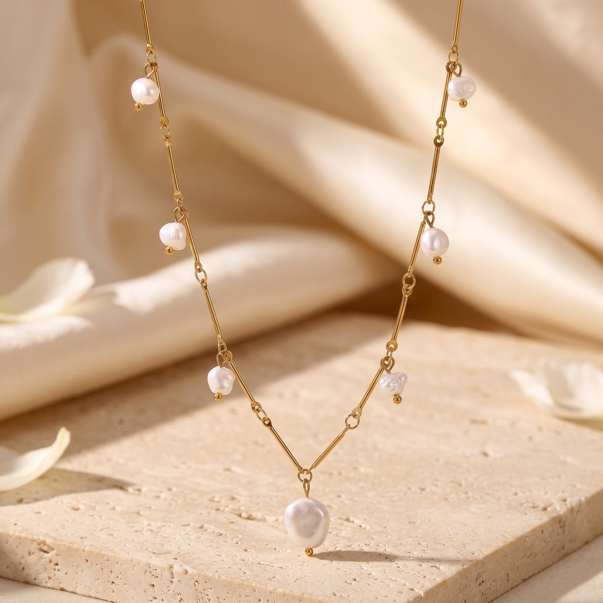 Gold finish stainless steel necklace with baroque freshwater pearls draped over a stone and silk background.